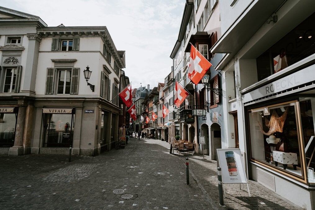 Image curtesy of Claudio Schwarz, Unsplash A street in Switzerland with Swiss flags hanging from shop fronts.
